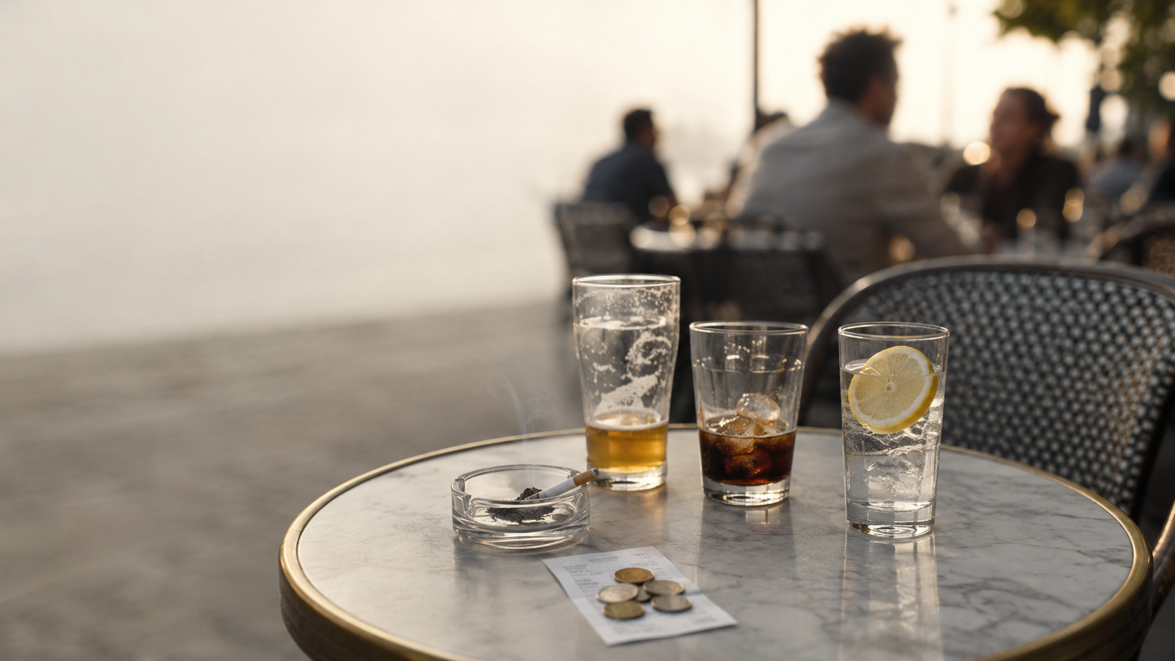 Taula d’una terrassa amb un cendrer i una cigarreta encesa, una cervesa a mig beure, un refresc de cola amb gel, un got d’aigua intacte amb llimona, un tiquet i unes monedes. Al fons, persones conversen desenfocades.